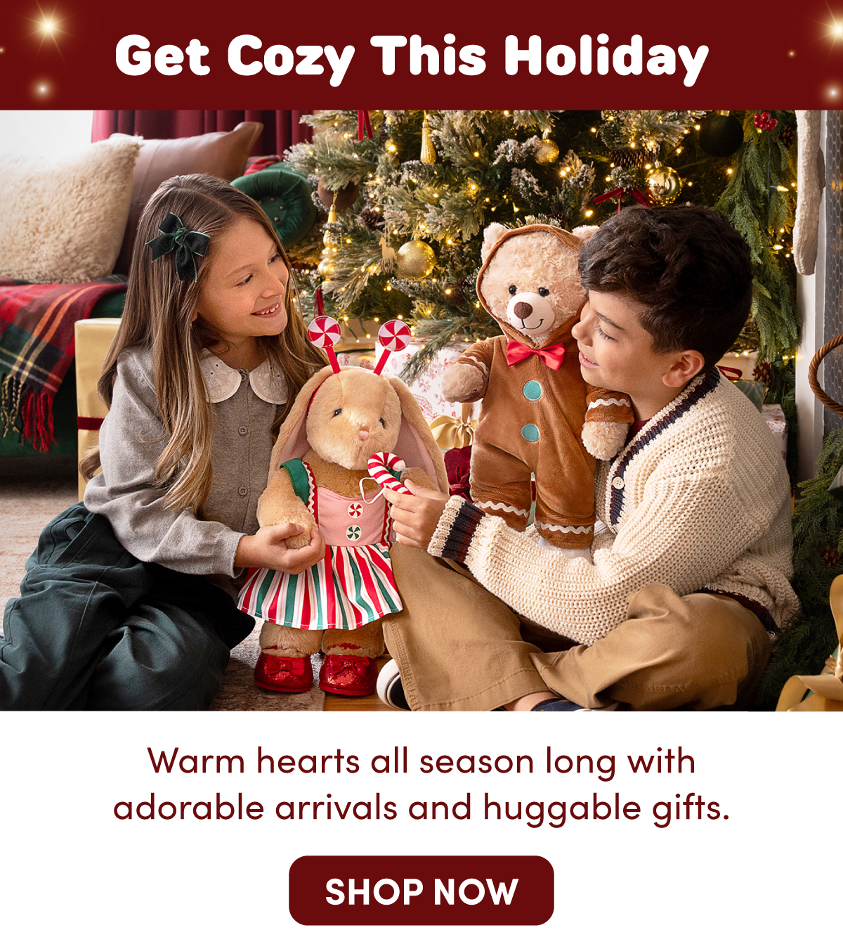 Get Cozy This Holiday. Two children sit beside a decorated Christmas tree, playing with plush gifts including a bunny dressed in a peppermint-inspired holiday outfit and a teddy bear dressed as a gingerbread cookie. Wrapped presents, glowing lights, gold ornaments, and festive décor surround the scene. Text reads: Get Cozy This Holiday. Warm hearts all season long with adorable arrivals and huggable gifts. SHOP NOW.