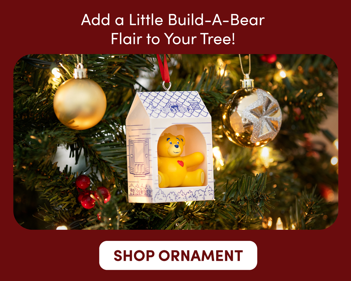Add a Little Build-A-Bear Flair to Your Tree! A Build-A-Bear holiday ornament hangs on a decorated Christmas tree, featuring a small yellow bear with a red heart displayed inside a cub condo-shaped ornament with blue illustrated details. Gold ornaments, twinkling lights, green pine branches, and red berries surround the scene. Text reads: Add a Little Build-A-Bear Flair to Your Tree! SHOP ORNAMENT.