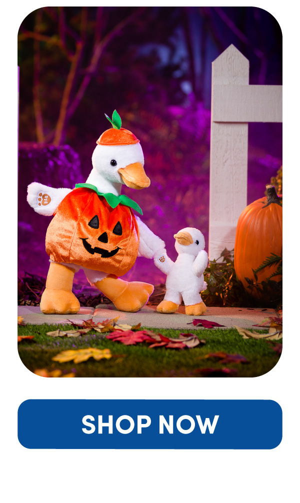 Regular-size Silly Goose plush dressed in a orange pumpkin costume with a matching pumpkin hat walks hand in hand with a Mini Beans Silly Goose plush. The pumpkin costume features a jack-o’-lantern face on the front. The pair are shown beside a white fence and pumpkin with fallen autumn leaves scattered along the path against a purple and pink Halloween backdrop.