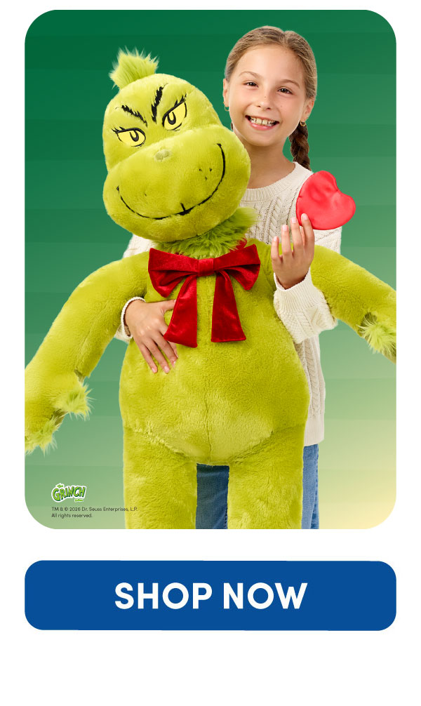 The Grinch™ graphic featuring a child holding an oversized Grinch plush with bright green fur, embroidered yellow eyes, a mischievous grin, and wearing a large red bow. The child also holds an oversized red heart-shaped accessory. The Grinch logo appears in the lower corner against a green gradient background
