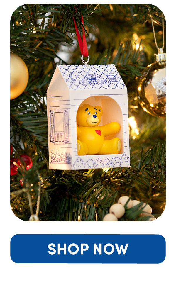 Decorative ornament hanging on a Christmas tree, featuring a small yellow teddy bear with a red heart sitting inside a cub condo. The cub condo is illustrated with blue line-art details including a window, shrubs, and a bear-themed emblem on the roof. Warm tree lights, gold ornaments, green pine branches, and red ribbon accents surround the ornament. No promotional text is shown.